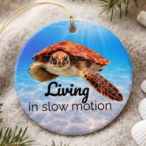 Peut inclure: Une décoration en céramique ronde illustrant une tortue marine nageant dans une eau bleue avec le texte Living in slow motion imprimé en noir. L'ornement comprend une ficelle dorée pour le suspendre et présente une image détaillée de la tortue.