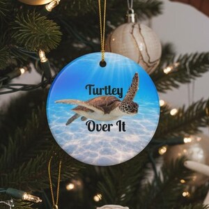 May include: A round ceramic Christmas ornament featuring a sea turtle swimming in clear blue ocean water. The text Turtley Over It is printed in black in the center. The ornament hangs from a gold string on a green pine tree branch.