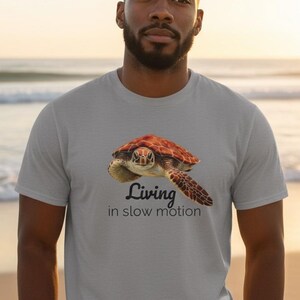 May include: A light grey short-sleeved t-shirt featuring a graphic of a brown sea turtle swimming. Below the turtle, the text reads "Living in slow motion" in a dark font. The shirt has a crew neckline and a standard fit.