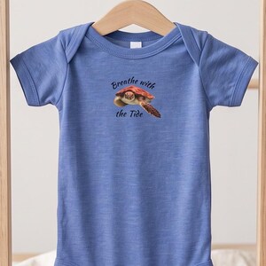 May include: A heather blue short-sleeved baby bodysuit featuring a graphic of a sea turtle and the text "Breathe with the Tide" printed in dark blue on the chest. The garment has a round neckline and a snap-button closure at the bottom.