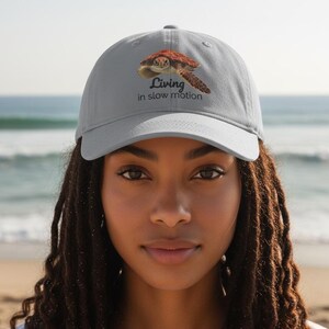 Puede incluir: Una gorra de béisbol gris claro con un gráfico de una tortuga marina marrón y el texto "Living in slow motion" impreso en negro. La gorra tiene una visera curva y una corona estructurada, ofreciendo un diseño inspirado en la naturaleza.