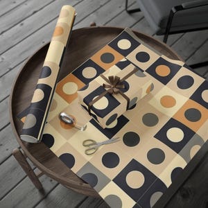 May include: A selection of gift wrapping items on a wooden table. A wrapped present with a brown ribbon sits on patterned wrapping paper. A roll of matching paper and scissors are also present. The paper has a geometric design with circles and squares in neutral tones.