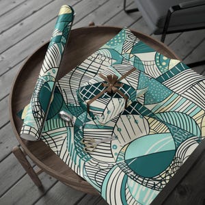 Könnte beinhalten: Eine Rolle Geschenkpapier mit Muster, ein verpacktes Geschenk und ein Blatt Papier mit ähnlichem Design. Das Design zeigt abstrakte Formen in Türkis, Weiß und Schwarz. Eine goldene Schere und ein silberner Klebebandabroller liegen ebenfalls auf dem Tisch.