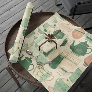 Pode incluir: Um rolo de papel de embrulho estampado e uma caixa de presente embrulhada com uma fita castanha estão sobre uma mesa de madeira. O papel apresenta um desenho repetido de bules e chávenas nas cores verde, pêssego e creme. Tesouras e fita adesiva também estão sobre a mesa.