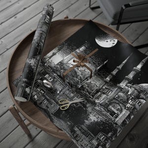 May include: Black and white wrapping paper with a detailed cityscape design, a wrapped gift with a brown ribbon, and a roll of matching paper. A pair of gold scissors and a silver ring are also on the wooden table.