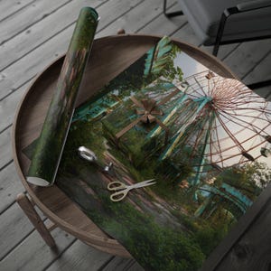 May include: Gift wrapping paper featuring a surreal image of an abandoned Ferris wheel and overgrown vegetation. A wrapped gift with a brown bow, scissors, and tape are on the paper. A roll of matching paper is also present.