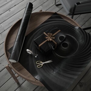 Peut inclure: Une boîte cadeau noire avec un ruban doré est posée sur du papier d'emballage noir à motif en spirale. Un rouleau de papier assorti, un distributeur de ruban adhésif et des ciseaux dorés sont sur une table en bois.