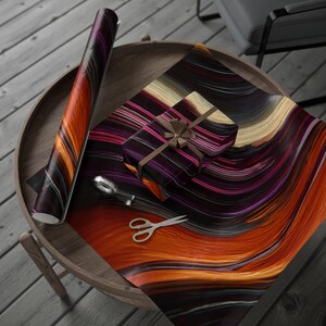 May include: A roll of wrapping paper and a wrapped gift with a brown ribbon sit on a wooden table. The paper features a colorful abstract design with orange, purple, and black streaks. Scissors and a tape dispenser are also on the table.