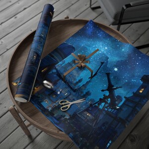 May include: A roll of blue wrapping paper and a wrapped gift with a brown ribbon sit on a wooden table. The wrapping paper and gift feature a dark blue cityscape design with a starry sky. Scissors and a silver tape dispenser are also on the table.