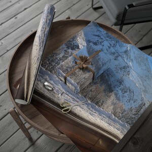 Puede incluir: Un regalo envuelto con un diseño de paisaje de montaña, atado con una cinta y lazo marrón, está sobre una mesa de madera. Papel de regalo a juego y tijeras doradas están cerca. La escena sugiere la entrega de regalos o la artesanía.