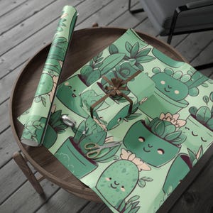 Puede incluir: Un rollo de papel de regalo verde y una caja de regalo envuelta con una cinta marrón, ambos con un patrón repetido de suculentas de dibujos animados en macetas. Un par de tijeras y una pequeña cuchara plateada también están sobre la mesa.