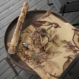 Puede incluir: Papel de regalo beige con diseño de pájaros y flores marrones. Un regalo envuelto está sobre la mesa, junto con unas tijeras y una cuchara. El papel está listo para usar.