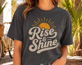Op de afbeelding: Een antracietgrijs t-shirt met korte mouwen en een afbeelding van een gele zon met stralen boven de tekst "Rise & Shine" in een crèmekleurig scriptlettertype. Het shirt heeft een ronde hals, een relaxte pasvorm en een verweerde print.