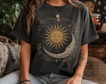 Op de afbeelding: Een antracietgrijs T-shirt met korte mouwen en een grafische print van een gouden zon en een maansikkel met gezichten, omringd door een cirkelvormig bloemenrankpatroon en kleine sterren. Het shirt heeft een ronde hals en opgerolde mouwboorden.