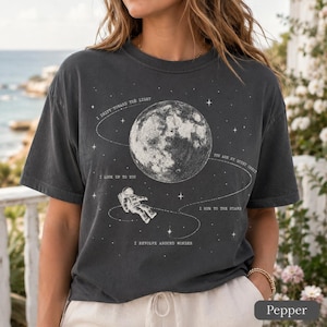 May include: A charcoal gray t-shirt featuring a white graphic of a moon, an astronaut, and stars. The text reads: I DRIFT TOWARD THE LIGHT, I LOOK UP TO YOU, YOU ARE MY QUIET ORBIT, I HUM TO THE STARS, and I REVOLVE AROUND WONDER with orbital lines.