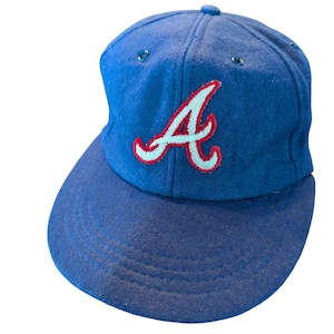 Könnte beinhalten: Blaue Baseballkappe mit gebogenem Schirm. Auf der Kappe ist ein großes, gesticktes "A" in Rot mit hellblauer Umrandung auf der Vorderseite zu sehen. Die Kappe ist aus einem weichen, filzartigen Material gefertigt.