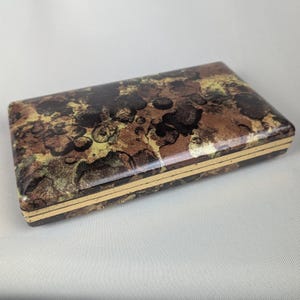 Peut inclure: Une boîte rectangulaire avec une finition brillante. La boîte présente un motif moucheté dans des tons de marron, noir et or. Les bords sont bordés d'une fine bande dorée, ajoutant une touche d'élégance au design.