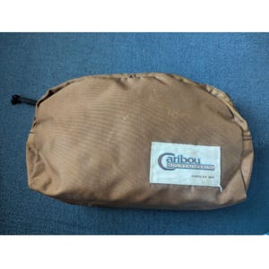 May include: A brown, semi-circular pouch with a black drawstring closure. A white rectangular label with the text "Caribou Mountaineering" is sewn on the front. The pouch is made of a durable, possibly water-resistant material.