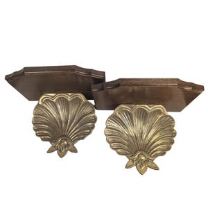 May include: A pair of wall-mounted shelves featuring dark brown wooden rectangular backings with scalloped edges and polished brass supports shaped like large, detailed seashells with prominent ridges and a decorative base element.