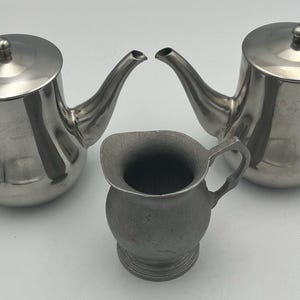 May include: Two polished stainless steel teapots with hinged lids, curved spouts, and black handles, accompanied by a smaller, matte-finish metal creamer pitcher with a wide pouring lip and a single handle, arranged together on a plain surface.