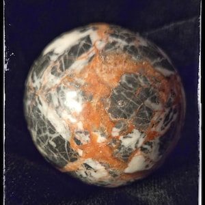 May include: A polished stone sphere with a marbled appearance. The sphere features a mix of colors, including white, gray, and orange, with intricate veining patterns. The stone is smooth and reflective, suggesting a decorative or collectible item.