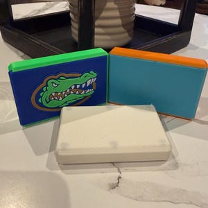 以下が含まれることがあります： 3つの長方形のケース。1つは緑のアリゲーターのグラフィックが入った青色、もう1つはオレンジ色のトップが付いたティール色、3つ目は白色です。ケースは白い表面にあります。
