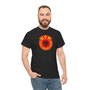 Może przedstawiać: Czarny t-shirt z czerwono-żółtą odznaką z napisem "UÇK" i słowami "Ushtria Çlirimtare e Kosovës" wokół dwugłowego orła. T-shirt jest noszony przez osobę w niebieskich dżinsach.