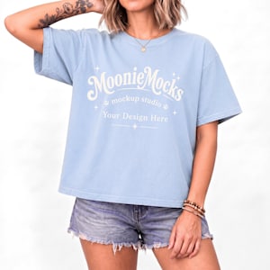 May include: A light blue short-sleeved t-shirt featuring white text that reads MoonieMocks mockup studio Your Design Here, accented with small star and paw print graphics. The shirt has a crew neckline and a relaxed, boxy fit.