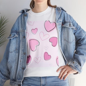 Puede incluir: Camiseta blanca con un estampado de corazones rosas con contornos negros sobre un fondo rosa claro. Los corazones varían en tamaño y están dispersos. Se usa con una chaqueta vaquera azul.