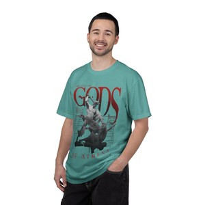 Puede incluir: Una camiseta de manga corta color verde azulado con un estampado gráfico de una estatua clásica en pose dinámica. El diseño incluye el texto rojo "GODS" y "OF ATHENA" junto con un texto negro más pequeño y tenue tras la ilustración.
