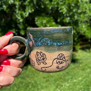 May include: Handmade ceramic mug with a dark teal and blue glaze. The mug features a brown base with black leaf designs. The handle is the same teal color as the rim. A unique piece for coffee or tea.