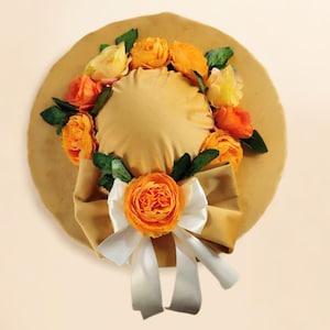 Può includere: Un cappello color beige con tesa larga, ornato di rose di tessuto arancioni e gialle e foglie verdi. Un grande fiocco bianco con un centro beige è sul davanti. Il cappello è un accessorio decorativo.
