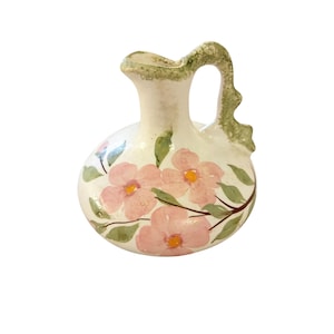 May include: A small ceramic pitcher with a rounded body and a single handle. The base color is off-white, decorated with hand-painted pink flowers featuring yellow centers, green leaves, and brown stems. The handle and rim have green accents.