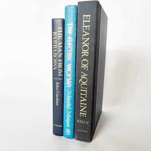 May include: Three hardcover books standing upright. The left book is dark blue with white text reading The Man From Barbarossa by John Gardner. The middle book is light blue with white text reading The Electric Woman by Marabel Morgan. The right book is black with gold text reading Eleanor of Aquitaine by Kelly.