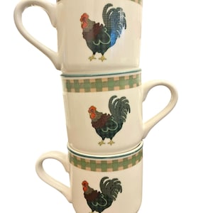 Puede incluir: Tres tazas de cerámica blanca apiladas con un diseño de gallo. Cada taza presenta una ilustración de gallo con acentos rojos, azules y verdes. Los bordes tienen un patrón de cuadros verdes y beige, y cada taza tiene un asa blanca.