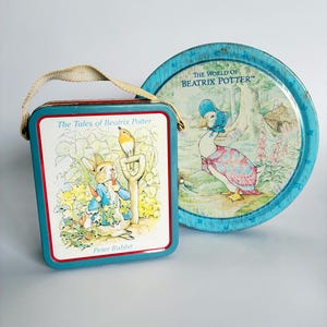 May include: A rectangular metal lunchbox featuring Peter Rabbit with the text The Tales of Beatrix Potter and a round metal tin featuring Jemima Puddle-Duck with the text The World of Beatrix Potter. Both containers have blue edges.