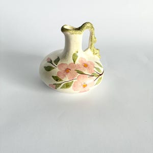 May include: A small, cream-colored ceramic pitcher featuring a hand-painted floral design with soft pink petals, yellow centers, and green leaves. The pitcher has a textured, moss-green handle and a matching green rim around the opening.
