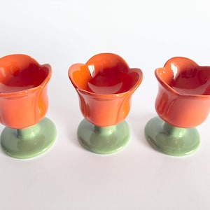 May include: Three ceramic egg cups shaped like tulips. Each cup features a glossy orange bowl with scalloped edges, supported by a smooth, light green circular base. The design mimics the appearance of a flower bloom on a short, sturdy stem.