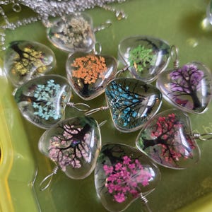 May include: A collection of heart-shaped pendants filled with colorful dried flowers. Each clear resin pendant features a different color scheme, including yellow, orange, blue, green, and pink. The pendants are attached to silver-colored chains.