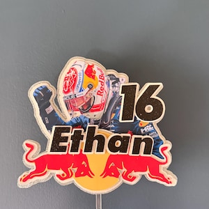 Puede incluir: Adorno para tarta con un piloto de carreras con casco, el número 16 y el nombre "Ethan". El logotipo de Red Bull es prominente. El adorno se sujeta con un palito de plástico transparente.