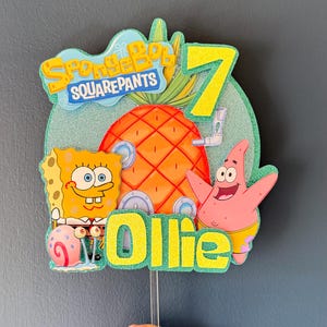 Op de afbeelding: Kleurrijke taarttopper met SpongeBob SquarePants, Patrick Star en een ananas. Het cijfer 7 en de naam "Ollie" zijn in glinsterend geel. De achtergrond is lichtblauw met glitter. Een verjaardagsdecoratie.