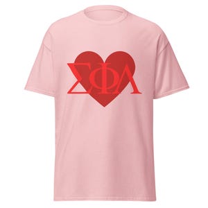 Op de afbeelding: Lichtroze T-shirt met een groot rood hart met de Griekse letters ΣΦΛ. Het t-shirt is gemaakt van zacht materiaal en heeft korte mouwen.