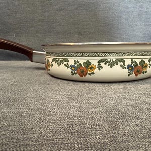May include: A vintage cream-colored saucepan with a floral pattern in orange, yellow, and blue. It has a brown handle and a silver rim. The pan is designed for cooking and is likely made of enamel or a similar material.