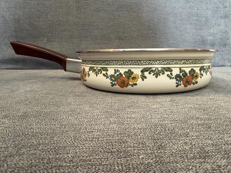 May include: A vintage cream-colored saucepan with a floral pattern in orange, yellow, and blue. It has a brown handle and a silver rim. The pan is designed for cooking and is likely made of enamel or a similar material.