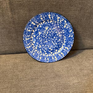 Może przedstawiać: Okrągły, niebiesko-biały ceramiczny talerz z czarną obwódką. Talerz ma nakrapiany wzór białych kropek na niebieskim tle. Talerz leży na szarej powierzchni.