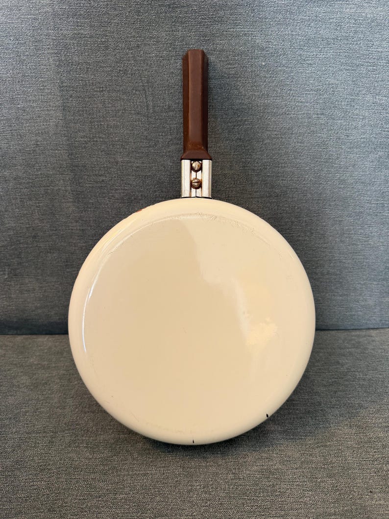 May include: A small, cream-colored frying pan with a dark brown handle. The pan has a rounded shape and a shiny surface. The handle is rectangular and attached to the pan with silver hardware. A vintage kitchenware item.