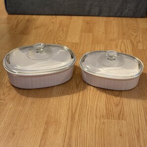 May include: Two white oval casserole dishes with glass lids. The dishes have a ribbed design and are of different sizes. The lids have a clear glass knob in the center. The dishes are on a wooden floor.