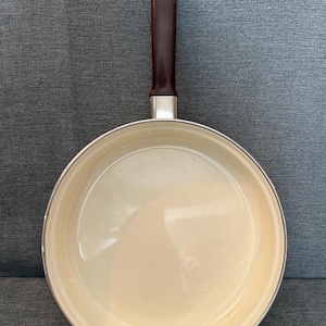 May include: A non-stick frying pan with a cream-colored interior and a dark brown handle. The handle is attached to the pan with a silver metal piece. The pan is round and has a slightly raised edge.