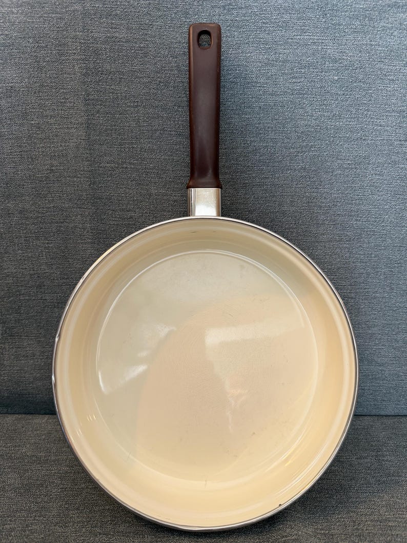 May include: A non-stick frying pan with a cream-colored interior and a dark brown handle. The handle is attached to the pan with a silver metal piece. The pan is round and has a slightly raised edge.
