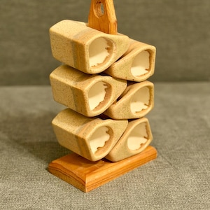 May include: A wooden wine glass rack with a natural finish. The rack holds six wine glasses in a stacked design. The rack is made of light brown wood and has a small wooden support at the top. The base is a rectangular shape.